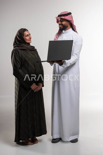 العمل الجماعي وروح الفريق الواحد، بورتريه لزميلين عربيين سعوديين خليجيين يقومان بمناقشة و تنظيم الاعمال بإستخدام جهاز الحاسوب المحمول، تبادل الأفكار والخبرات لتحقيق أهداف الشركة، دمج العمل مع التقنية والتكنولوجيا، صورة الجسم بالطول الكامل، خلفية بيضاء.