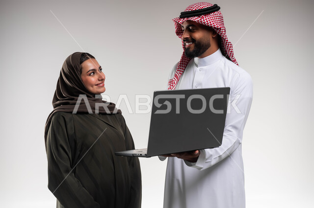 تبادل الأفكار والخبرات لتطوير استراتيجيات العمل، بورتريه مقرب لزميلين عربيين خليجيين سعوديين مبتسمين يقومان بإنجاز المهام عبر الحاسوب المحمول، التعاون وتوحيد الجهود لتحقيق أهداف الشركة، دمج التقنية و التكنولوجيا في العمل، خلفيه بيضاء