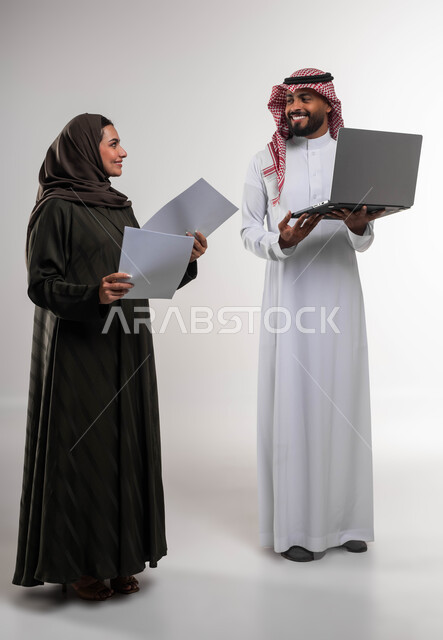 تدريب الموظفين على الأساليب الجديدة في العمل لتحقيق أهداف الشركة، دمج العمل مع التقنية والتكنولوجيا، بورتريه لزميلين عربيين خليجيين سعوديين مبتسمين يقومان بإنجاز المهام عبر الحاسوب المحمول، العمل الجماعي وروح الفريق الواحد، صورة الجسم بالكامل، خلفيه بيضاء