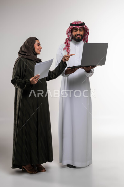 مناقشة المهام و تحقيق أهداف الشركة، بورتريه لزميلين عربيين خليجيين سعوديين مبتسمين يقومان بإنجاز المهام عبر الحاسوب المحمول، الاشارة الى اللابتوب، التعاون وروح الفريق الواحد، تعليم الموظفين أساليب عمل جديدة، صورة الجسم بالكامل، خلفيه بيضاء