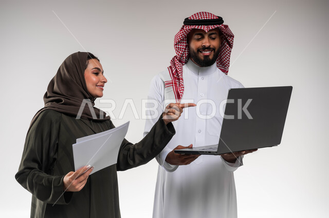 الاحترافية في أداء المهام للتطور المهني و تحقيق الأهداف، دمج العمل مع التقنية والتكنولوجيا، بورتريه مقرب لزميلين عربيين خليجيين سعوديين مبتسمين يقومان بإنجاز المهام عبر الحاسوب المحمول، الاشارة الى اللابتوب، العمل الجماعي و روح الفريق الواحد، خلفيه بيضاء