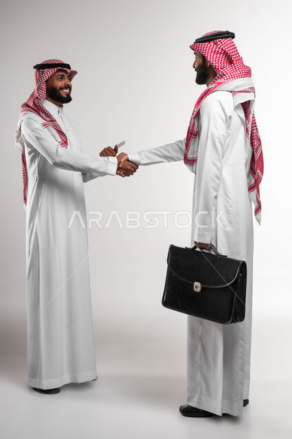 Partnership and agreement on business and commercial projects, portrait of two smiling Gulf Arab Saudi businessmen wearing traditional Saudi dress, shaking hands and concluding new business deals, discussing tasks to achieve the organization’s goals, getting to know each other and building social relationships, full body photo, white background