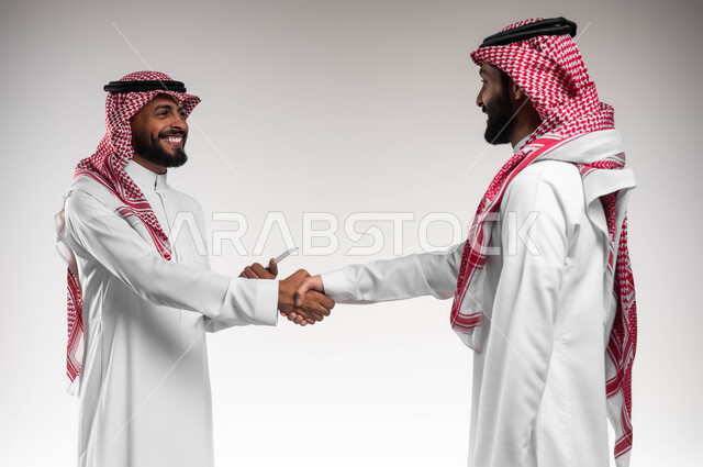 الإتفاق والشراكة على الأعمال والمشاريع التجارية، بورتريه مقرب لرجلي اعمال عربيين سعوديين خليجين مبتسمين يرتديان الزي السعودي التقليدي يقومان بالمصافحة وعقد صفقات عمل جديدة، مناقشة المهام لتحقيق أهداف المؤسسة، التعارف وبناء العلاقات الاجتماعية، خلفية بيضاء