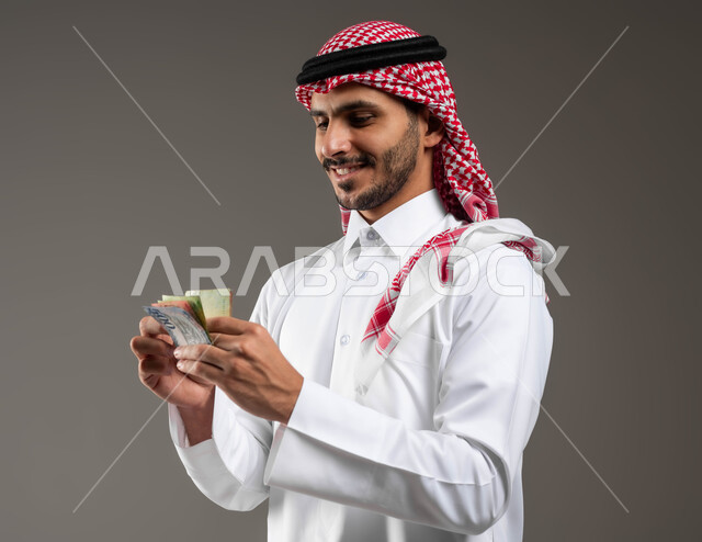 العملات النقدية والورقية في المملكة, إدراة المشاريع الاقتصادية, بورتريه مقرب لرجل عربي خليجي سعودي مبتسم يرتدي الثوب التقليدي والشماغ يحمل أوراق النقود من الريال السعودي, مفهوم الوفرة المالية, خلفية رمادية
