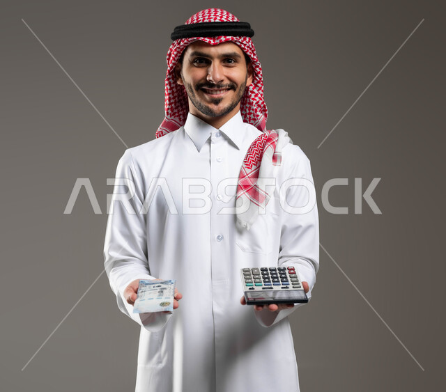 الأمور الاقتصادية والمشاريع المستقبلية, مفهوم ادرة الشؤون الاقتصادية, بورتريه مقرب لرجل عربي خليجي سعودي يرتدي الثوب التقليدي والشماغ يحمل في يده عملة ورقية للريال السعودي وآلة حاسبة في اليد الاخرى, استخدام الالات الرقمية لإجراء العمليات الحسابية, خلفية رمادية