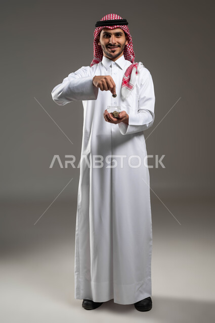 Standing and looking at the camera with happy gestures, the concept of savings and financial abundance, financing future projects and businesses, portrait of a smiling Saudi Gulf Arab man wearing a traditional thobe and shemagh collecting money in a glass bowl, full-length body portrait, gray background