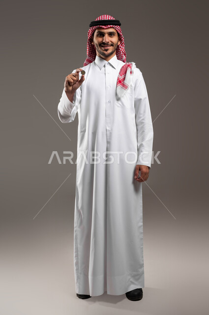 النمو والأرباح المالية, الاستثمار في قطاع الاقتصاد السعودي, توفير الأموال ومفهوم الإدخار, بورتريه لرجل عربي خليجي سعودي مبتسم يرتدي الثوب التقليدي والشماغ ينظر إلى الكاميرا ويحمل بيده عملة نقدية للريال السعودي, صورة الجسم بالطول الكامل, خلفية رمادية