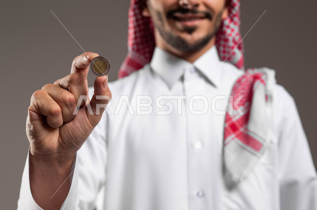الاستثمار في قطاع الاقتصاد السعودي, توفير الأموال ومفهوم الإدخار, بورتريه مقرب لرجل عربي خليجي سعودي مبتسم يرتدي الثوب التقليدي والشماغ ينظر إلى الكاميرا ويحمل بيده عملة نقدية للريال السعودي, النمو والأرباح المالية, خلفية رمادية