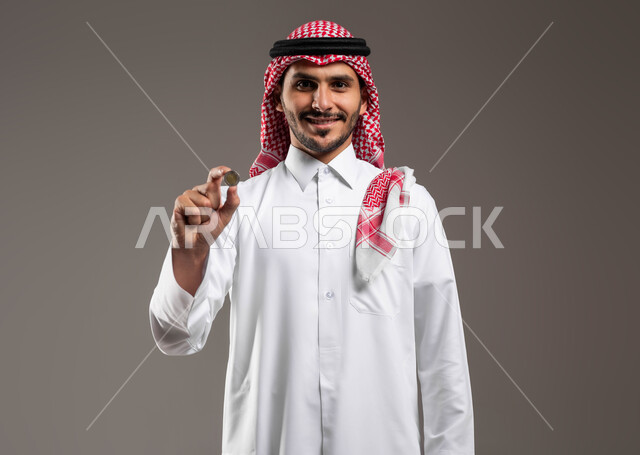 توفير الأموال ومفهوم الإدخار, بورتريه مقرب لرجل عربي خليجي سعودي مبتسم يرتدي الثوب التقليدي والشماغ ينظر إلى الكاميرا ويحمل بيده عملة نقدية للريال السعودي, الاستثمار في قطاع الاقتصاد السعودي, النمو والأرباح المالية, خلفية رمادية
