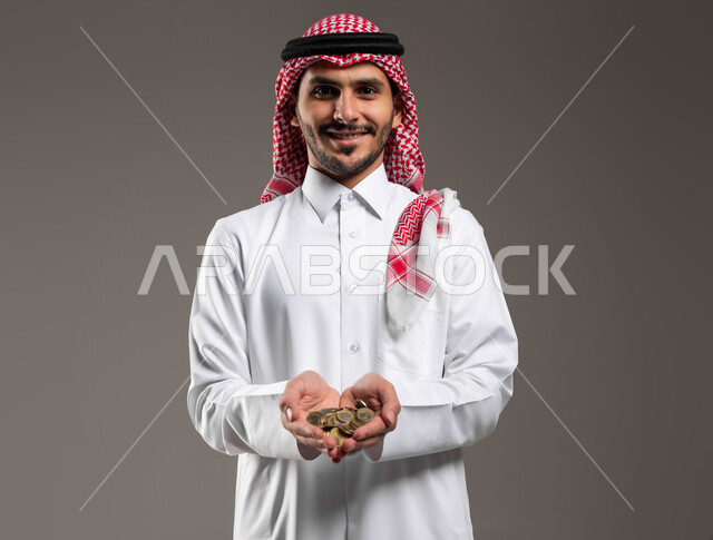 الفوائد والاستثمار في المشاريع المستقبلية, مفهوم تمويل الأعمال، بورتريه مقرب لرجل عربي خليجي سعودي مبتسم يرتدي الثوب التقليدي والشماغ ينظر إلى الكاميرا ويحمل بيده كومة من العملات المعدنية، النمو والأرباح المالية، إدخار وتوفير الاموال, خلفية رمادية