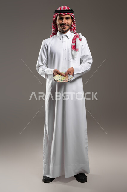 الاستثمار بالمشاريع الاقتصادية, الوقوف بإيماءات السعادة بالربح المالي الوفير, بورتريه لرجل عربي خليجي سعودي مبتسم يرتدي الثوب التقليدي والشماغ يمسك بيده أموال نقدية من الريال السعودي, إدارة الحسابات والقروض البنكية, صورة الجسم بالكامل, خلفية رمادية