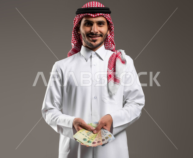الوقوف بإيماءات السعادة بالربح المالي الوفير, العملات الورقية في المملكة, الاستثمار بالمشاريع الاقتصادية, بورتريه لرجل عربي خليجي سعودي مبتسم يرتدي الثوب التقليدي والشماغ يمسك بيده أموال نقدية من الريال السعودي, إدارة الحسابات والقروض البنكية, خلفية رمادية