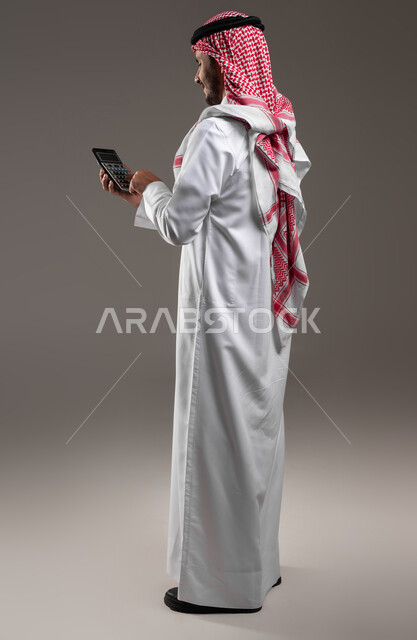 حساب التكاليف والأرباح والخسائر, تنظيم الشؤون المالية, بورتريه من الخلف لرجل عربي خليجي سعودي يرتدي الثوب التقليدي والشماغ يقوم باستخدام الآلة الحاسبة لإجراء العمليات الحسابية, الاستثمار في القطاع الاقتصادي في السعودية, صورة الجسم بالطول الكامل, خلفية رمادية