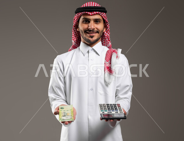 مفهوم إدارة الشؤون الاقتصادية, الوقوف باستقامة والنظر إلى الكاميرا ببشاشة, بورتريه لرجل عربي خليجي سعودي يرتدي الثوب التقليدي والشماغ يحمل في يده عملة ورقية للريال السعودي وآلة حاسبة في اليد الاخرى, استخدام الالات الرقمية لإجراء العمليات الحسابية، خلفية رمادية