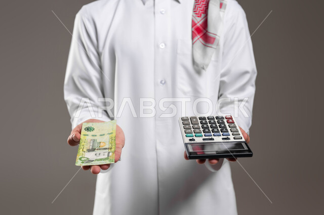 الأمور الاقتصادية والمشاريع المستقبلية, مفهوم ادرة الشؤون الاقتصادية, بورتريه لرجل عربي خليجي سعودي يرتدي الثوب التقليدي والشماغ يحمل في يده عملة ورقية للريال السعودي وآلة حاسبة في اليد الاخرى, استخدام الالات الرقمية لإجراء العمليات الحسابية, خلفية رمادية