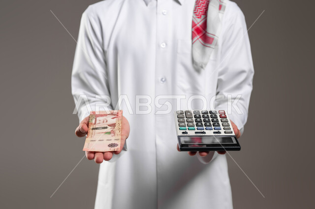 الاستثمار والمشاريع المستقبلية, بورتريه لرجل عربي خليجي سعودي يرتدي الثوب التقليدي والشماغ يحمل في يده عملة ورقية للريال السعودي وآلة حاسبة في اليد الاخرى, استخدام الالات الرقمية لإجراء العمليات الحسابية، مفهوم ادرة الشؤون الاقتصادية, خلفية رمادية