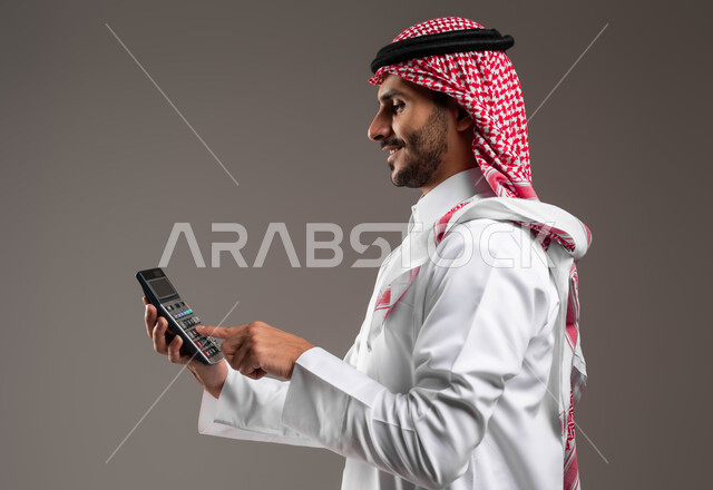 العمل في إدارة الحسابات, بورتريه من الجانب لرجل عربي خليجي سعودي يرتدي الثوب التقليدي والشماغ يقوم باستخدام الآلة الحاسبة لإجراء العمليات الحسابية, حساب التكاليف والأرباح والخسائر, تنظيم الشؤون المالية, الاستثمار في القطاع الاقتصادي في السعودية, خلفية رمادية