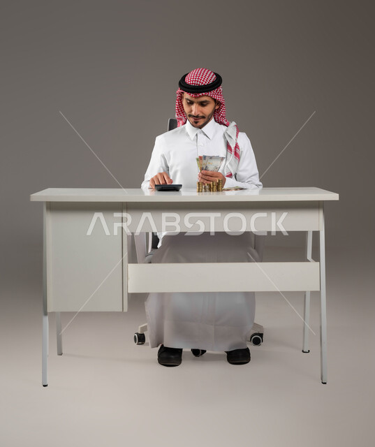 بيئة العمل المكتبية, الاستثمار الاقتصادي والمعاملات المالية, استخدام الآلة الحاسبة لإجراء العمليات الحسابية, حسابات القروض البنكية, بورتريه لرجل عربي خليجي سعودي يرتدي الثوب التقليدي والشماغ يمسك بيده النقود الورقية ويعمل في إدارة الحسابات, خلفية رمادية