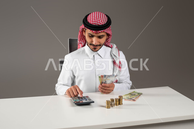 الاستثمار الاقتصادي والمعاملات المالية, استخدام الآلة الحاسبة لإجراء العمليات الحسابية, حسابات القروض البنكية, تقديم خدمات الرواتب للموظفين, بورتريه مقرب لرجل عربي خليجي سعودي يرتدي الزي التقليدي يمسك بيده النقود الورقية ويعمل في إدارة الحسابات, خلفية رمادية