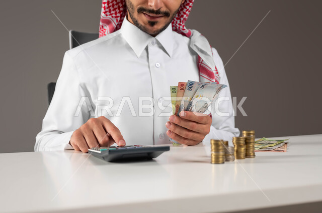 استخدام الآلة الحاسبة لإجراء العمليات الحسابية, حسابات القروض البنكية, المعاملات المالية والاستثمار الاقتصادي, تقديم خدمات الرواتب للموظفين, بورتريه مقرب لرجل عربي خليجي سعودي يرتدي الزي التقليدي يمسك بيده النقود الورقية ويعمل في إدارة الحسابات, خلفية رمادية