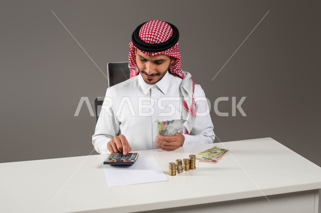 Financial transactions and economic investment, providing payroll services to employees, portrait of a Saudi Gulf Arab man wearing traditional clothing working in accounts management, using a calculator to perform calculations, bank loan invoices and accounts, office work environment, gray background