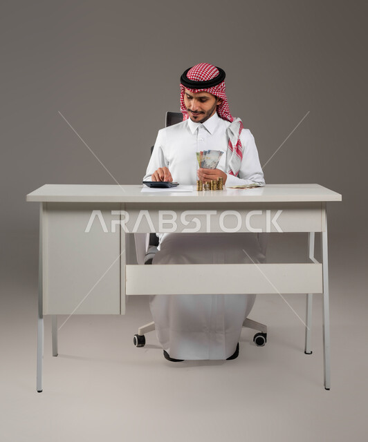 تقديم خدمات الرواتب للموظفين, بورتريه لرجل عربي خليجي سعودي يرتدي الزي التقليدي يعمل في إدارة الحسابات, استخدام الآلة الحاسبة لإجراء العمليات الحسابية, فوايتر وحسابات القروض البنكية, بيئة العمل المكتبية, المعاملات المالية والاستثمار الاقتصادي, خلفية رمادية