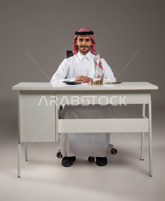 إدارة شؤون العمل الاقتصادية, الجلوس بثقة والنظر إلى الكاميرا بسعادة, تسديد الفواتير المالية, تقديم الخدمات وتحويل الأموال, الاستثمار في قطاع الاقتصاد السعودي, بورتريه لرجل عربي خليجي سعودي يرتدي الزي التقليدي يعمل بمجال الحسابات, خلفية رمادية