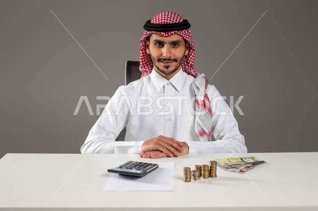 الجلوس بثقة والنظر إلى الكاميرا بسرور, تسديد الفواتير المالية, تقديم الخدمات وتحويل الأموال, الاستثمار في قطاع الاقتصاد السعودي, بورتريه مقرب لرجل عربي خليجي سعودي يرتدي الزي التقليدي يعمل بمجال الحسابات, إدارة شؤون العمل الاقتصادية, خلفية رمادية