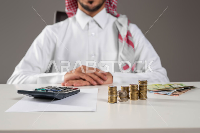 تسديد الفواتير المالية, تقديم الخدمات وتحويل الأموال, الاستثمار في قطاع الاقتصاد السعودي, بورتريه مقرب لرجل عربي خليجي سعودي يرتدي الزي التقليدي يجلس بثقة ويعمل بمجال الحسابات, إدارة شؤون العمل الاقتصادية, خلفية رمادية