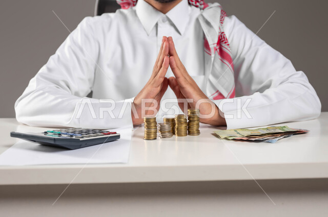 تقديم الخدمات وتحويل الأموال, الاستثمار في قطاع الاقتصاد السعودي, بورتريه مقرب لرجل عربي خليجي سعودي يرتدي الزي التقليدي  يضع يديه بشكل هرمي على الطاولة ويعمل بمجال الحسابات, إدارة شؤون العمل الاقتصادية, تسديد الفواتير المالية, خلفية رمادية