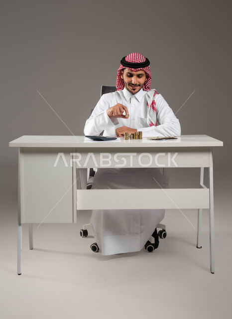 بيئة العمل المكتبية, تمويل المشاريع الاستثمارية, إدخار وتوفير المال, الحسابات والقروض البنكية, مهن ووظائف سعودية, بورتريه لرجل عربي خليجي سعودي يرتدي الثوب التقليدي والشماغ يمسك بيده عملة الريال المعدنية, خلفية رمادية