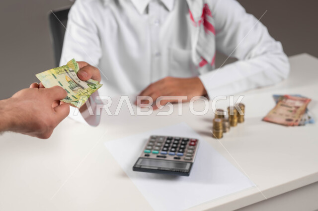 العمل في إدارة الحسابات, الإحصائيات والقروض البنكية, بيئة العمل المكتبية, بورتريه لرجل عربي خليجي سعودي يرتدي الثوب التقليدي والشماغ يقوم بمسك عملة ورقية من فئة الخمسين ريال, مفهوم الوفرة المالية, التعامل بالعملات النقدية والورقية المعدنية, خلفية رمادية
