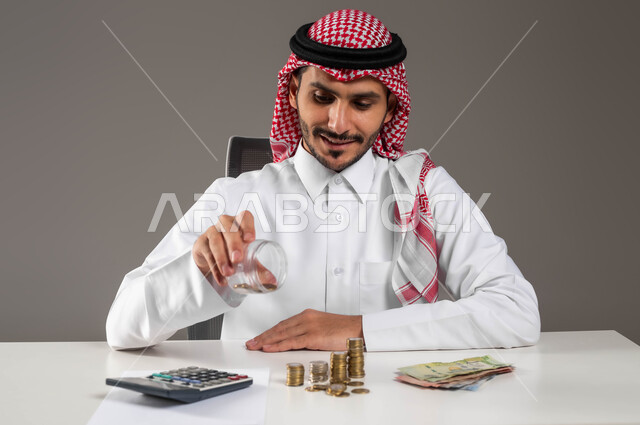 إدراة الشؤون الاقتصادية, مفهوم الاستدامة المالية, توفير المال لبناء المشاريع والخطط المستقبلية, بورتريه مقرب لرجل عربي خليجي سعودي مبتسم يرتدي الثوب التقليدي والشماغ يفرغ النقود من الوعاء الزجاجي, عملة الريال الورقية والمعدنية في المملكة, خلفية رمادية