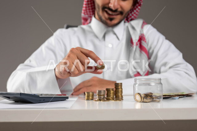 الإحصائيات والحسابات البنكية, النهضة والنمو الاقتصادي في المملكة, التعامل بالعملات الورقية والمعدنية في السعودية, بورتريه لرجل عربي خليجي سعودي مبتسم يرتدي الثوب التقليدي والشماغ يمسك بيده قطعة نقود بإيماءات السرور, مفهوم الوفرة المالية, خلفية رمادية