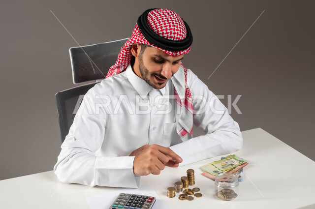 النهضة والنمو الاقتصادي في المملكة, التعامل بالعملات الورقية والمعدنية في السعودية, بورتريه لرجل عربي خليجي سعودي مبتسم يرتدي الثوب التقليدي والشماغ يمسك بيده قطعة نقود بإيماءات السرور, مفهوم الوفرة المالية, الإحصائيات والحسابات البنكية, خلفية رمادية