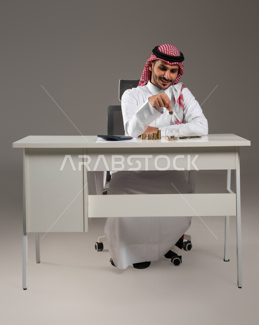 تسديد الحسابات والقروض البنكية, إدراة شؤون العمل المالية والمكتبية, توفير المال لبناء المشاريع المسقبلية, بورتريه لرجل عربي خليجي سعودي مبتسم يرتدي الثوب التقليدي والشماغ يقوم بإدخار الأموال, مفهوم الاقتصاد والاستثمار المالي, خلفية رمادية