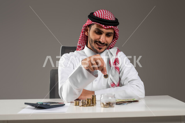 إدراة شؤون العمل المالية والمكتبية, توفير المال لبناء المشاريع المسقبلية, تسديد الحسابات والقروض البنكية, بورتريه لرجل عربي خليجي سعودي مبتسم يرتدي الثوب التقليدي والشماغ يقوم بإدخار الأموال, مفهوم الاقتصاد والاستثمار المالي, خلفية رمادية