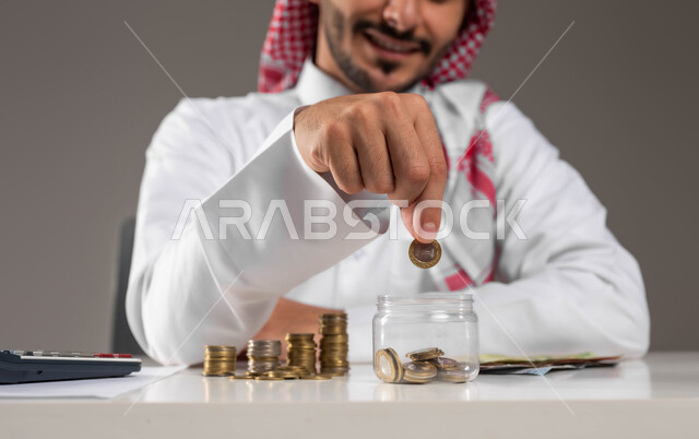 توفير المال لبناء المشاريع المسقبلية, تسديد الحسابات والقروض البنكية, إدراة شؤون العمل المالية والمكتبية, بورتريه لرجل عربي خليجي سعودي مبتسم يرتدي الثوب التقليدي والشماغ يقوم بإدخار الأموال, مفهوم الاقتصاد والاستثمار المالي, خلفية رمادية