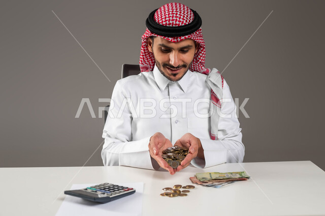 بيئة العمل المكتبية, إدخار وتوفير الأموال، بورتريه لرجل عربي خليجي سعودي مبتسم يرتدي الثوب التقليدي والشماغ يحمل بيده عملات نقدية معدنية للريال السعودي، استخدام الالة الحاسبة لاجراء العمليات الحسابية, مفهوم الاقتصاد والاستثمار المالي, خلفية رمادية