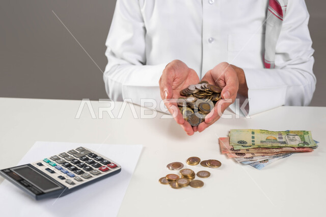 مفهوم الاقتصاد والاستثمار المالي, بيئة العمل المكتبية, إدخار وتوفير الأموال، بورتريه لرجل عربي خليجي سعودي يرتدي الثوب التقليدي يحمل بيده عملات نقدية معدنية للريال السعودي، استخدام الالة الحاسبة لاجراء العمليات الحسابية, خلفية رمادية