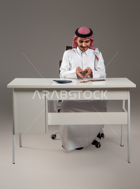 بيئة العمل المكتبية, إدخار وتوفير الأموال، بورتريه لرجل عربي خليجي سعودي يرتدي الثوب التقليدي والشماغ يحمل بيده عملات نقدية معدنية للريال السعودي، استخدام الالة الحاسبة لاجراء العمليات الحسابية, مفهوم الاقتصاد والاستثمار المالي, خلفية رمادية