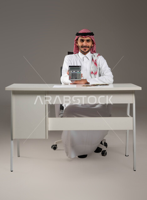 الحسابات والتمويل الاقتصادي للمشاريع المستقبلية, توفير ودراسة الأمور المالية, إجراء العمليات الحسابية باستخدام الآلة الحاسبة الرقمية, تسديد حساب القروض البنكية, بورتريه لرجل عربي خليجي سعودي مبتسم يرتدي الثوب التقليدي والشماغ ينظر إلى الكاميرا, خلفية رمادية