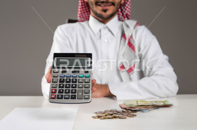 إجراء العمليات الحسابية, توفير ودراسة الأمور المالية,  الحسابات والتمويل الاقتصادي للمشاريع المستقبلية, تسديد حساب القروض البنكية, بورتريه لرجل عربي خليجي سعودي مبتسم يرتدي الثوب التقليدي والشماغ يمسك بيده الآلة الحاسبة الرقمية, خلفية رمادية