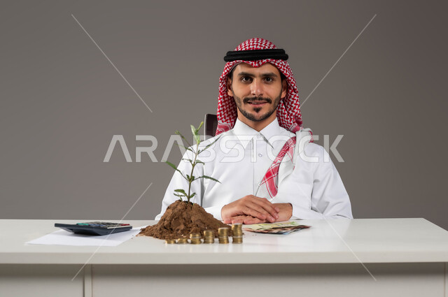 النمو والنهضة الاقتصادية في المملكة, التعامل بالعملات الورقية والمعدنية في السعودية, بورتريه لرجل عربي خليجي سعودي مبتسم يرتدي الثوب التقليدي والشماغ ينظر إلى الكاميرا بإيماءات السرور, مفهوم الوفرة المالية, الإحصائيات والحسابات البنكية, خلفية رمادية