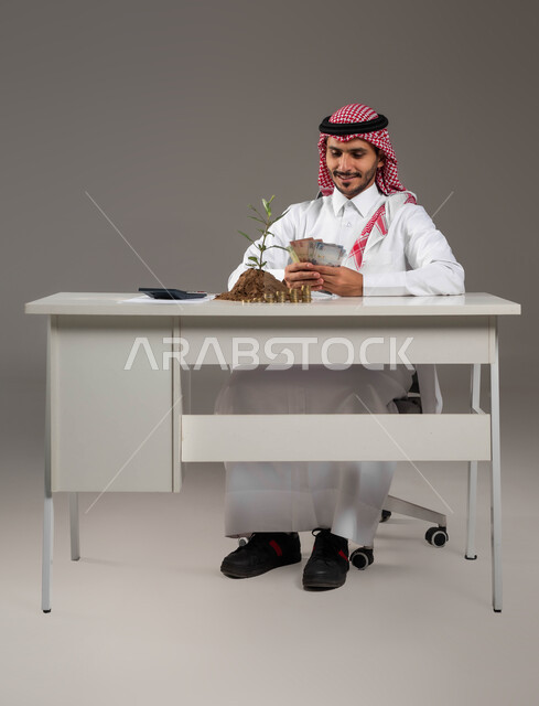 النمو والأرباح المالية, إيماءات السرور بتمويل المشاريع المستقبلية, تنظيم أمور الحسابات والقروض البنكية, بورتريه لرجل عربي خليجي سعودي مبتسم يرتدي الثوب التقليدي والشماغ يمسك بيده عملة الريال الورقية, الاستثمار في القطاع الاقتصادي, خلفية رمادية