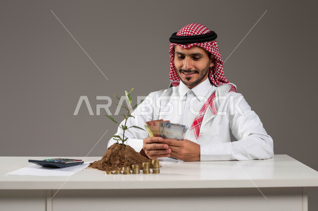 إيماءات السرور بتمويل المشاريع المستقبلية, تنظيم أمور الحسابات والقروض البنكية, بورتريه لرجل عربي خليجي سعودي مبتسم يرتدي الثوب التقليدي والشماغ يمسك بيده عملة الريال الورقية, النمو والأرباح المالية, الاستثمار في القطاع الاقتصادي, خلفية رمادية