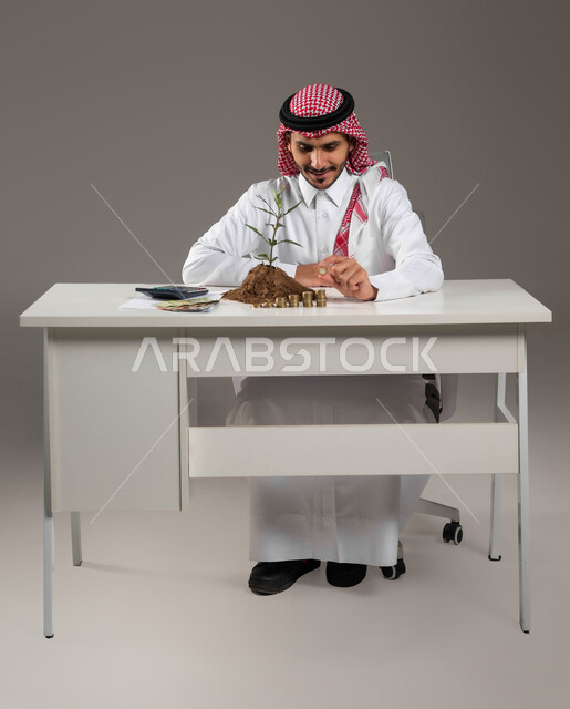 تنظيم الحسابات والقروض البنكية, تمويل المشاريع المستقبلية, مفهوم الاستثمار الاقتصادي, التوفير والإدخار والتخزين المالي, بيئة العمل المكتبية, بورتريه لرجل عربي خليجي سعودي مبتسم يرتدي الثوب التقليدي والشماغ يمسك بيده عملة معدنية, خلفية رمادية