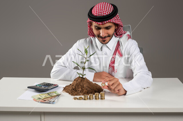 تمويل المشاريع المستقبلية, مفهوم الاستثمار الاقتصادي, ترتيب وتنظيم الحسابات والقروض البنكية, التوفير والإدخار والتخزين المالي, بيئة العمل المكتبية, بورتريه لرجل عربي خليجي سعودي مبتسم يرتدي الثوب التقليدي والشماغ يمسك بيده عملة معدنية, خلفية رمادية