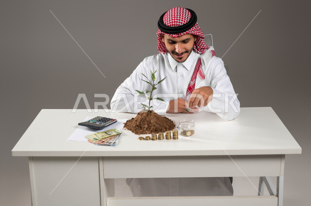 التوفير والإدخار والتخزين المالي, بيئة العمل المكتبية, مفهوم الاستثمار الاقتصادي, تمويل المشاريع المستقبلية, بورتريه لرجل عربي خليجي سعودي يرتدي الثوب التقليدي والشماغ يقوم بإحصاء الأموال, ترتيب وتنظيم الحسابات والقروض البنكية, خلفية رمادية