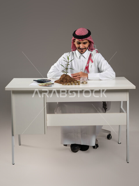 مفهوم الوفرة المالية, التخطيط في تمويل المشاريع المستقبلية, بورتريه لرجل عربي خليجي سعودي مبتسم يرتدي الثوب التقليدي والشماغ يجلس في مكتبه بإيماءات السرور والسعادة بالإدخار المالي, الاستثمار في الاقتصاد السعودي, خلفية رمادية